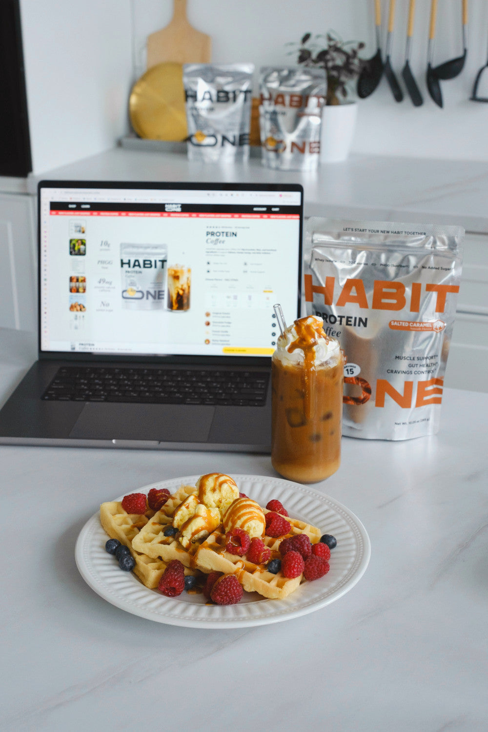 Iced HabitONE Protein Coffee next to waffles, a laptop and the bag